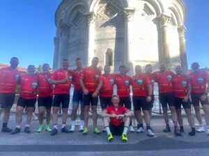 Group of cyclists in red jerseys posing together in front of a large historic stone monument with arches and columns against a bright blue sky.